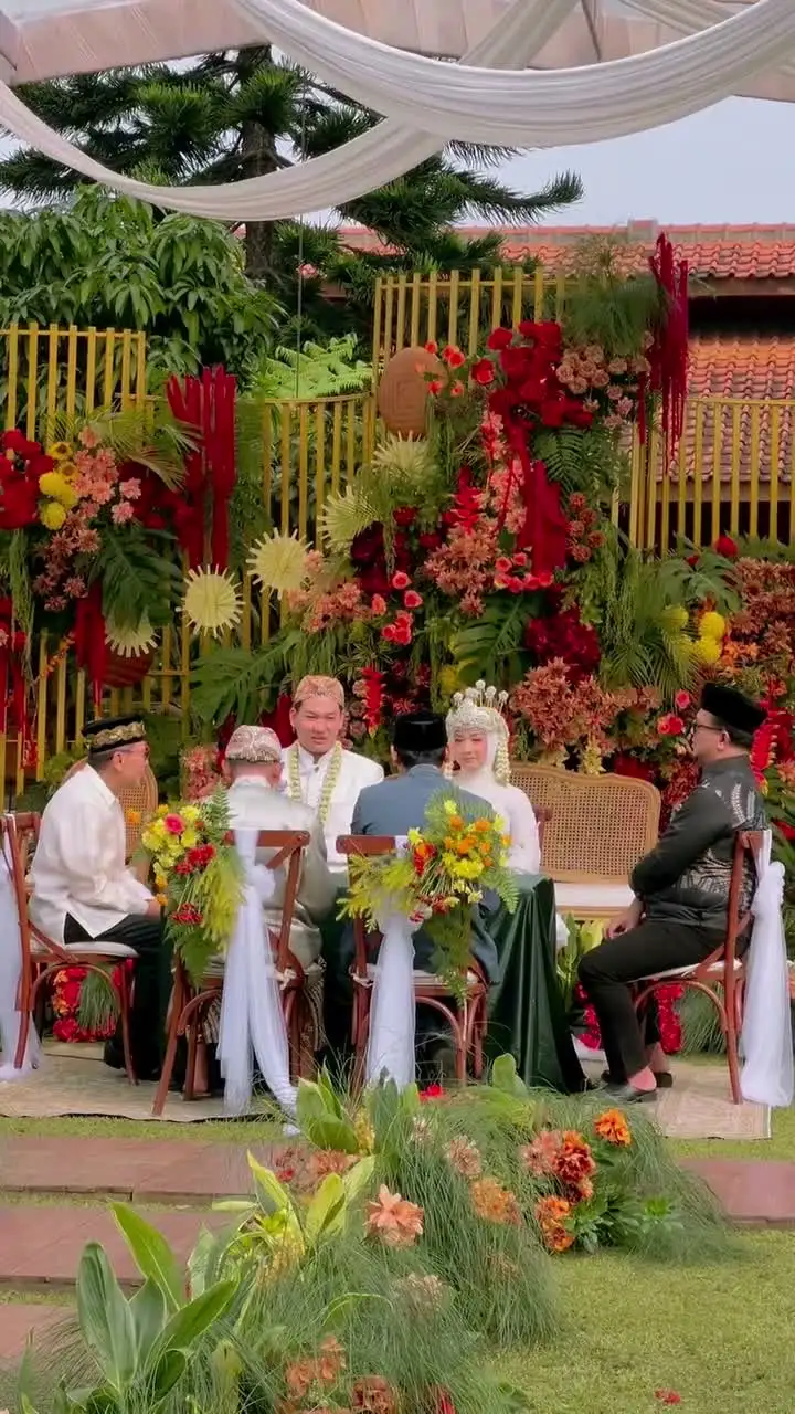 A newly married couple walking across the lawn under a gold ceremonial umbrella as guests look on