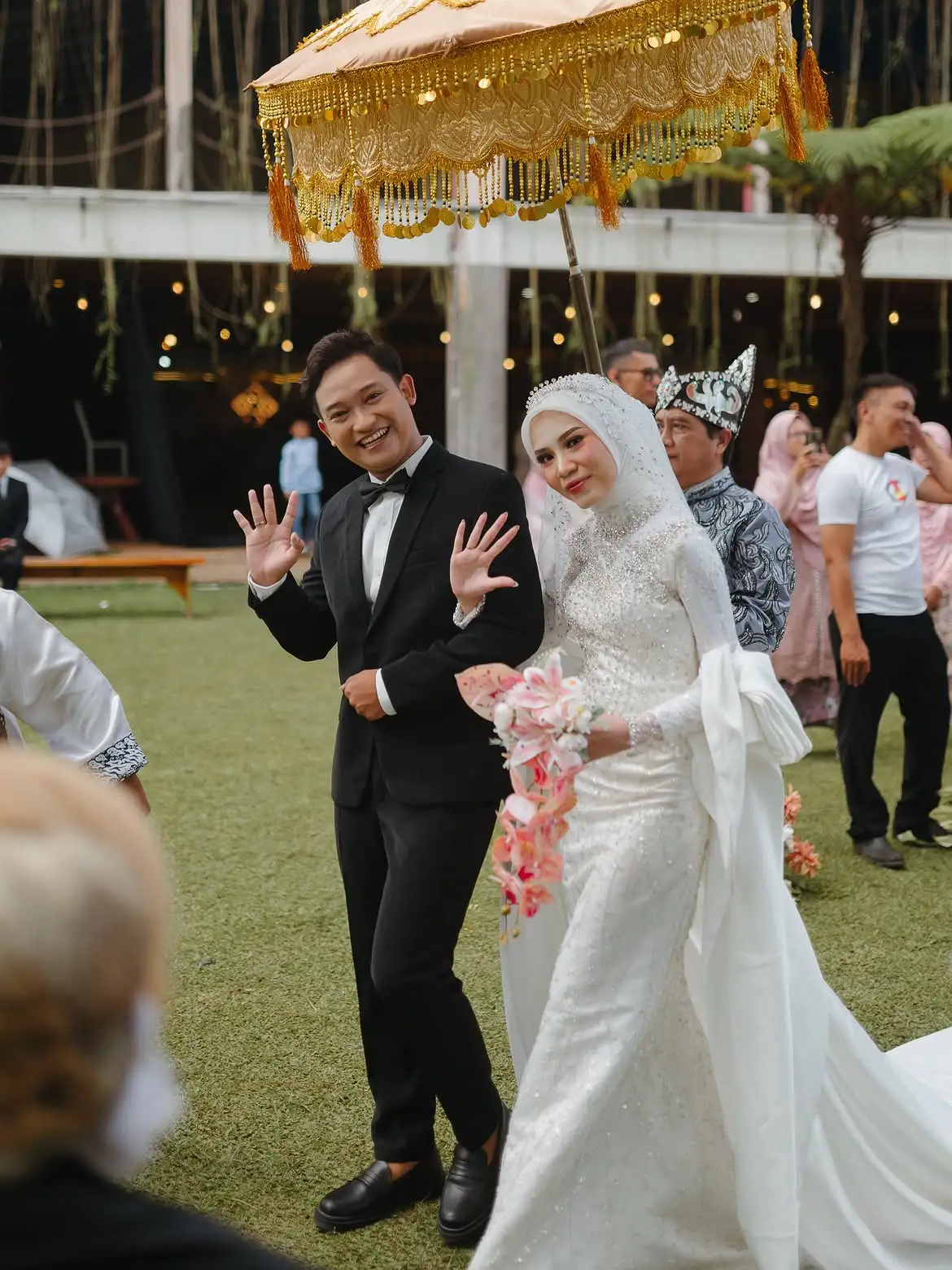 The couple walking in under a gold ceremonial umbrella as guests look on